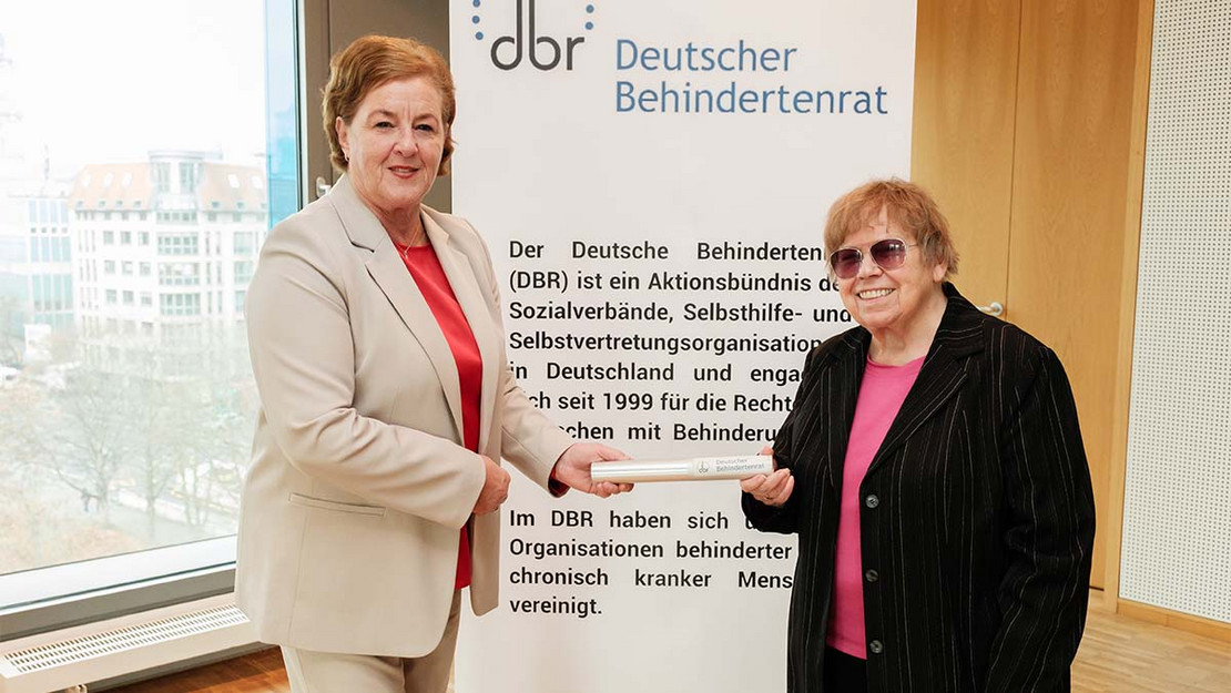 Eine Frau übergibt einer anderen einen Staffelstab. Im Hintergrund ein Aufsteller des Deutschen Behindertenrats. 