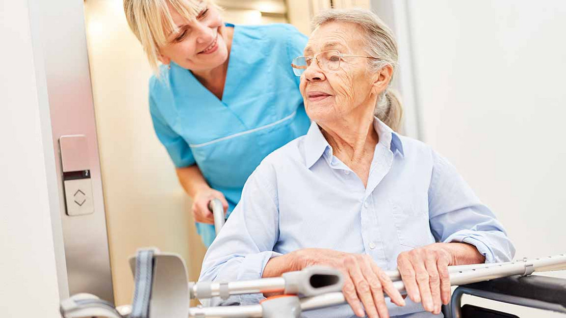 Pflegekraft schiebt eine Seniorin im Rollstuhl in einen Aufzug. 
