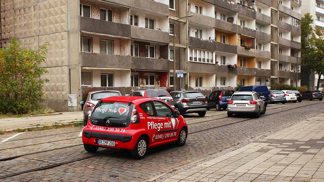 Auto eines Pflegediensts fährt durch ein Plattenbaugebiet. 