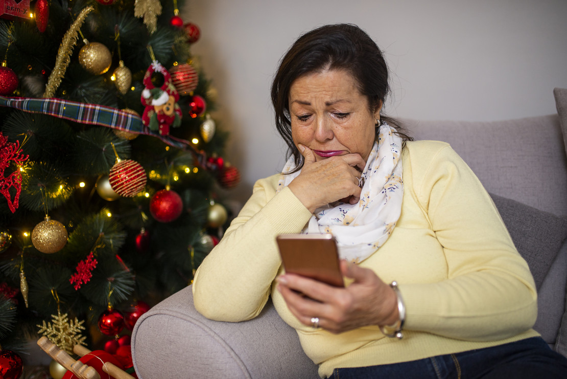Frau sitzt mit Handy in der Hand vor einem Weihnachtsbaum. 