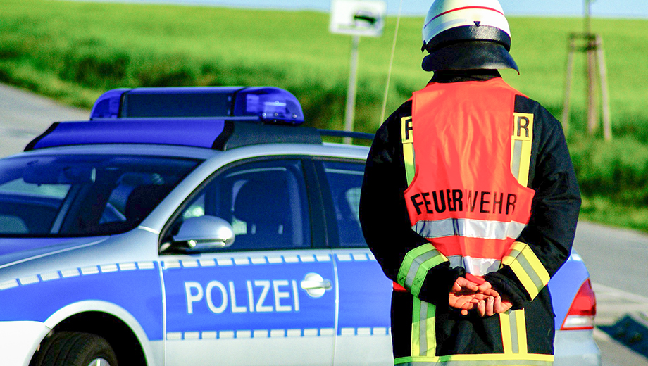 Bild eines Feuerwehrmannes vor einem Polizeiauto. Der Mann ist von hinten zu sehen.