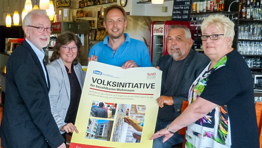 Mitglieder des SoVD halten ein Plakat für die Volksinitiative für bezahlbaren Wohnraum in der Hand.