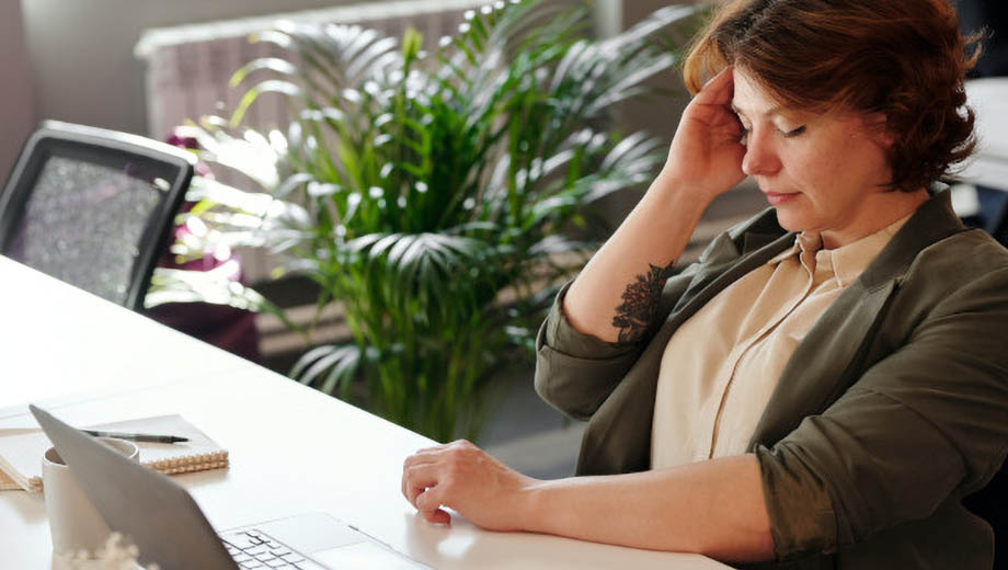 Ein Frau sitzt an ihrem Schreibtisch vor ihrem Laptop. Sie hält sich eine Hand an den Kopf und wirkt niedergeschlagen.
